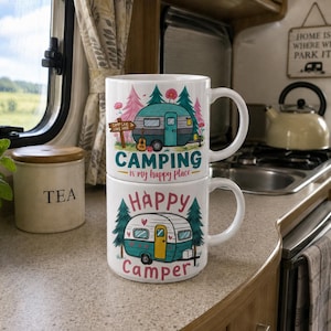 May include: Two white ceramic mugs stacked. The top mug features a teal camper design with the text "Camping is my happy place." The bottom mug has a teal camper design with the text "Happy Camper."