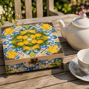May include: A square wooden box featuring a vibrant pattern of yellow lemons, green leaves, and blue and white floral tile motifs. The box has a hinged lid, a metallic latch, and four ornate bronze-coloured feet on the bottom corners.