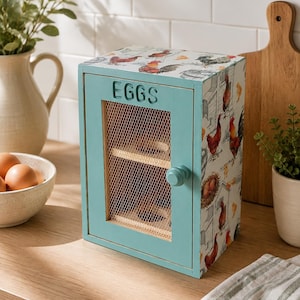 May include: A teal wooden egg storage cabinet with a wire mesh door, a round knob, and the word EGGS printed on the front. The sides and top feature a decorative pattern of roosters, hens, and nests. The interior contains two wooden shelves.
