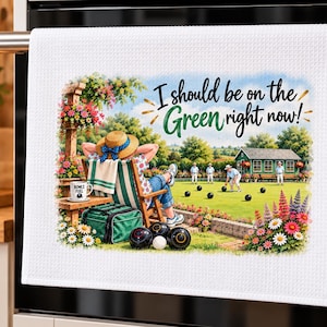 May include: A white waffle-weave tea towel featuring a colourful illustration of a person relaxing in a lawn chair watching a lawn bowls game. The text reads, I should be on the Green right now! A green bag, black bowls, and flowers are visible.