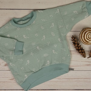 Peut inclure: Un pull bébé vert clair avec un motif d'oiseaux et de feuilles blancs. Le pull a des manches longues et un col rond.