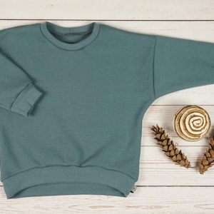 Puede incluir: Una sudadera de punto de gofre de color verde azulado con cuello redondo y mangas largas. Los puños y el dobladillo son acanalados. La camisa se exhibe sobre una superficie de madera clara, con piñas decorativas y una espiral de madera.