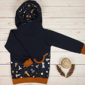 May include: A navy blue hooded sweatshirt with a space-themed pattern. The hood lining and lower front panel feature a print of rockets, planets, and stars. The cuffs and hem are a contrasting brown color. The sweatshirt is designed for children.