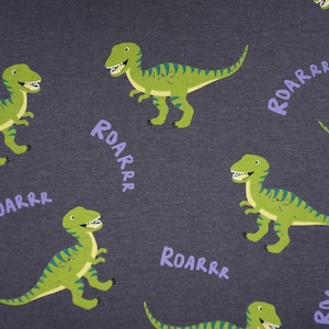 Può includere: Tessuto grigio scuro con un motivo ripetuto di dinosauri verdi e la parola "ROARRR" in viola. I dinosauri hanno strisce verdi e blu.