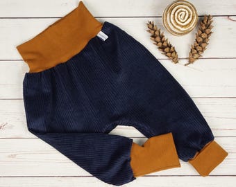 May include: Navy blue corduroy pants with a wide, mustard-colored waistband and cuffs. The pants are designed with a relaxed fit and are displayed on a light wood-grain surface. Decorative pinecones and a wooden spiral are also visible.