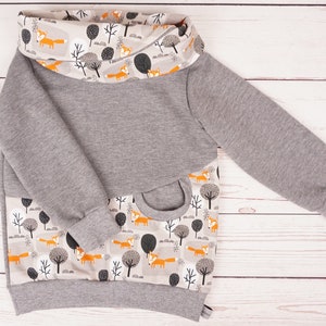 Könnte beinhalten: Ein grauer Pullover mit einem Fuchs- und Baumdruck am unteren Rand und einem passenden Druck am Rollkragen.