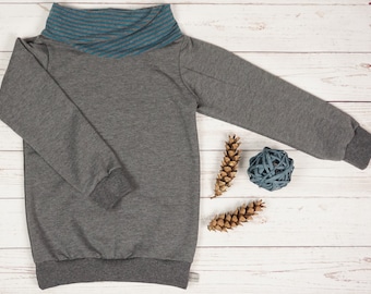 May include: A gray long-sleeved sweater with a blue and gray striped turtleneck collar. The sweater is laying on a white wooden surface with two pine cones and a ball of blue yarn.