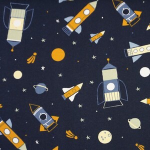Könnte beinhalten: Ein marineblauer Stoff mit einem sich wiederholenden Muster aus gelben und blauen Raketen, Sternen und Planeten. Die Raketen sind in verschiedenen Stilen, einige mit Fenstern und andere mit Flammen.