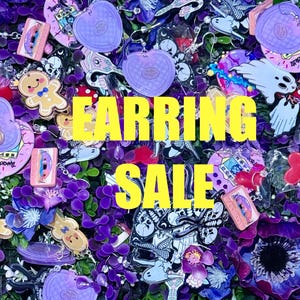 May include: Earring sale! A collection of colourful earrings featuring diverse designs such as hearts, ghosts, gingerbread men, and skulls. The earrings are presented on a bed of purple flowers and foliage. The text "EARRING SALE" is in yellow.