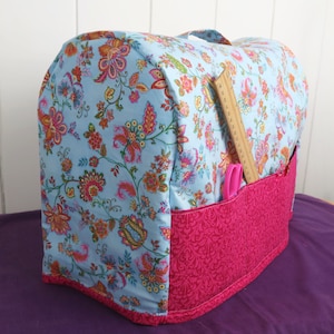Puede incluir: Una funda para m&aacute;quina de coser floral azul y rosa con un bolsillo para guardar herramientas de costura. La funda tiene un forro de tela rosa y una regla de madera sobresale del bolsillo.