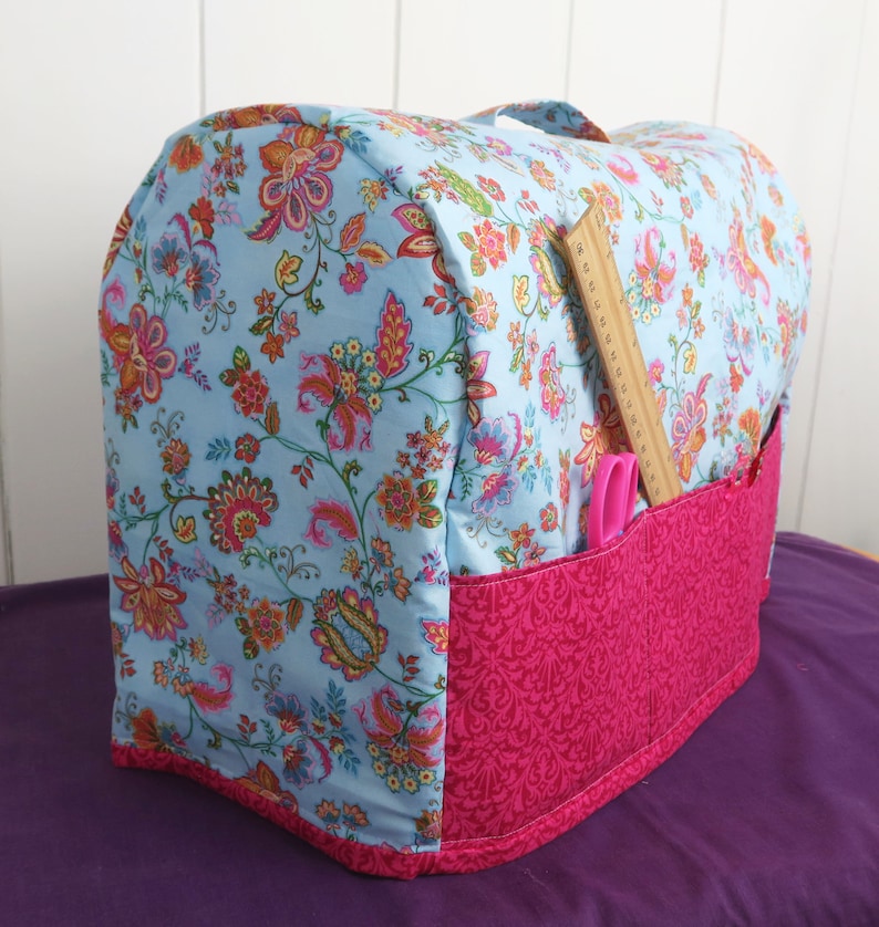 Puede incluir: Una funda para m&aacute;quina de coser floral azul y rosa con un bolsillo para guardar herramientas de costura. La funda tiene un forro de tela rosa y una regla de madera sobresale del bolsillo.