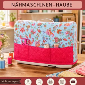 Könnte beinhalten: Eine Nähmaschinenhaube aus hellblauem Stoff mit buntem Blumenmuster und einem einfarbigen, dunkelrosa Taschenbereich. Die Haube hat einen Tragegriff und ist mit einer rosa Schere sowie Garnrollen in den Vordertaschen abgebildet.