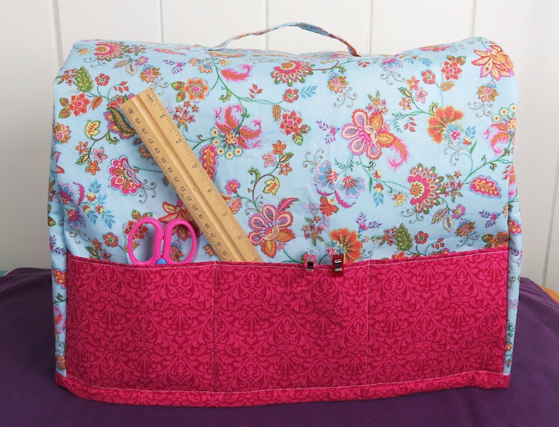 Puede incluir: Una funda para m&aacute;quina de coser de flores azules y rosas con un bolsillo de tela rosa y una regla y unas tijeras en el interior.
