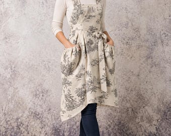 May include: A beige apron featuring a black toile de jouy pattern with pastoral scenes, wide shoulder straps, a tie waist, and two large front pockets. The apron is worn over a long-sleeved cream shirt, dark blue jeans, and black ankle boots.
