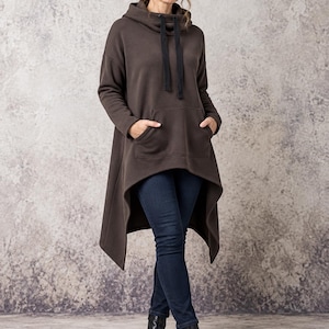 May include: A dark brown long-sleeve hooded tunic featuring a high-low hemline, a front kangaroo pocket, and black drawstring cords. The garment is paired with dark blue denim jeans and black ankle boots.