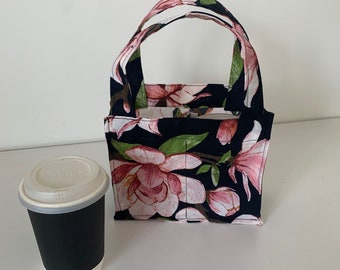 May include: A small black canvas tote bag with a floral print featuring pink flowers on a dark background. The bag has a single handle and is designed to carry a coffee cup.