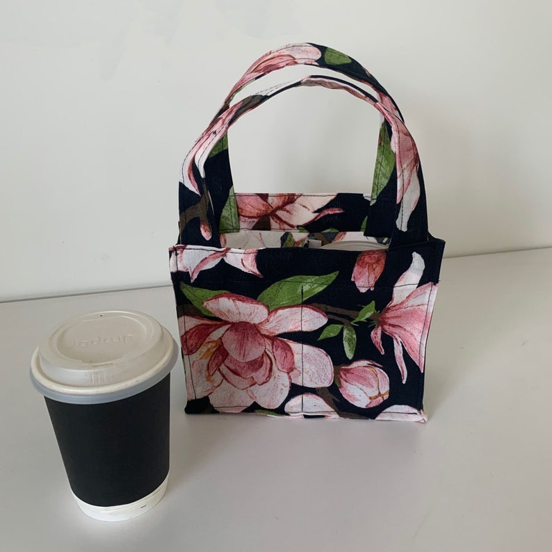 May include: A small black canvas tote bag with a floral print featuring pink flowers on a dark background. The bag has a single handle and is designed to carry a coffee cup.
