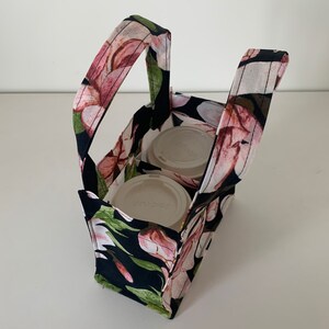 May include: A black fabric tote bag with a floral print featuring pink flowers and green leaves. The bag has two handles and two compartments for carrying two to-go coffee cups. The cups are white with black lids and the text "Sapporo" is visible on one of the cups.