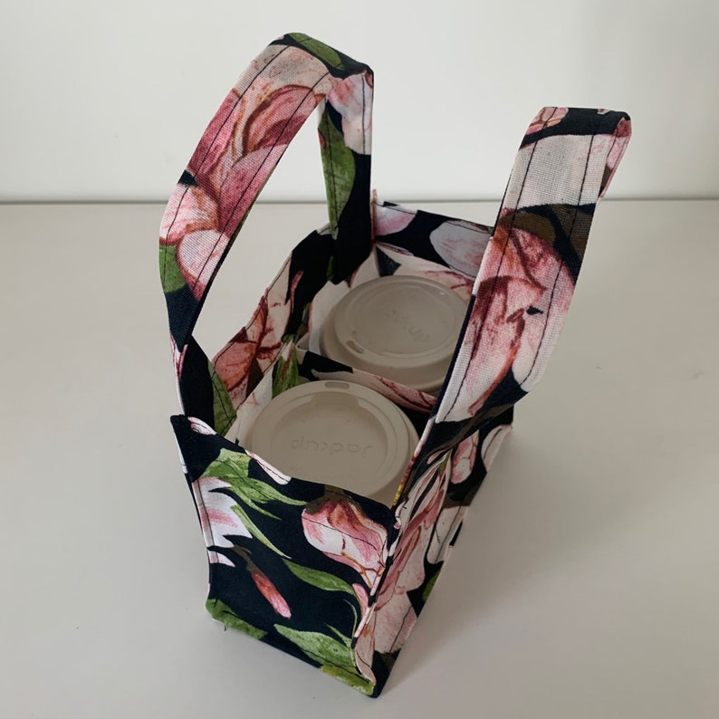 May include: A black fabric tote bag with a floral print featuring pink flowers and green leaves. The bag has two handles and two compartments for carrying two to-go coffee cups. The cups are white with black lids and the text "Sapporo" is visible on one of the cups.