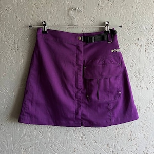 Könnte beinhalten: Ein lilafarbener Minirock mit einer großen Cargo-Tasche auf der rechten Seite. Der Rock hat einen schwarzen Gürtel mit einer silbernen Schnalle und ein Columbia Sportswear-Logo auf der Rückseite.