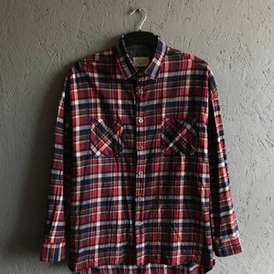May include: A red, white, and blue plaid flannel shirt with long sleeves and two chest pockets. The shirt is hanging on a black hanger against a gray textured wall.