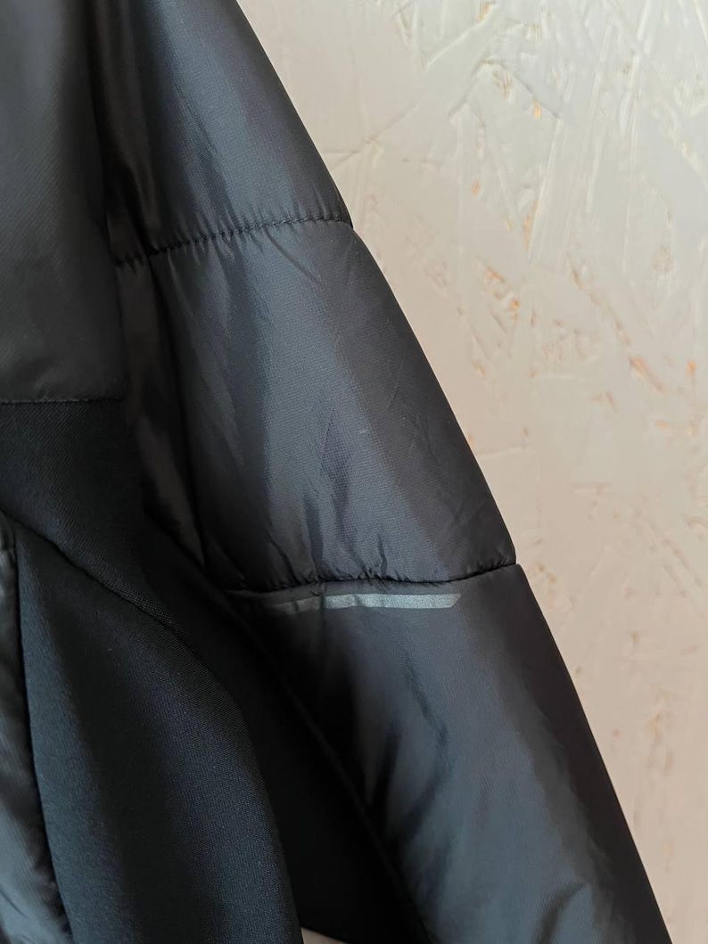 May include: Close-up of a black jacket with a quilted design. The jacket has a smooth, shiny texture and a reflective strip detail. The garment appears to be made of a lightweight material.
