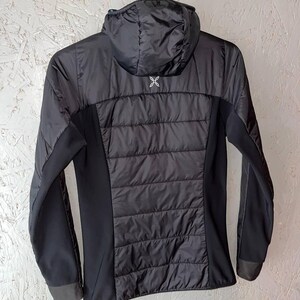 May include: Black hooded jacket with a quilted back panel and side panels. The jacket has long sleeves and a small white logo on the upper back. The jacket is hanging on a metal hanger.