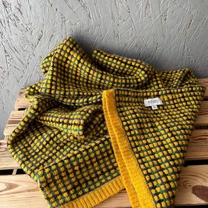 May include: A knitted scarf featuring a textured grid pattern in yellow, green, and black. The scarf has a solid yellow ribbed border along the edge and a white fabric label sewn onto the interior that reads KENZO PARIS with the letter U.