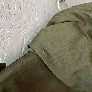 K&ouml;nnte beinhalten: Eine olivgr&uuml;ne Jacke mit glatter Stoffstruktur und einem sichtbaren kleinen Riss oder losen Faden im Bereich der Schulternaht. Das Kleidungsst&uuml;ck ist mit einem strukturierten Kragen versehen und h&auml;ngt an einem Metallhaken.