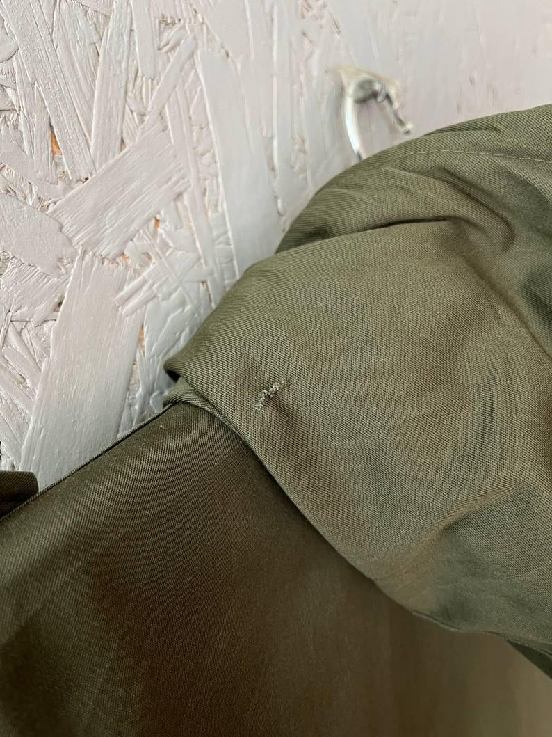 K&ouml;nnte beinhalten: Eine olivgr&uuml;ne Jacke mit glatter Stoffstruktur und einem sichtbaren kleinen Riss oder losen Faden im Bereich der Schulternaht. Das Kleidungsst&uuml;ck ist mit einem strukturierten Kragen versehen und h&auml;ngt an einem Metallhaken.