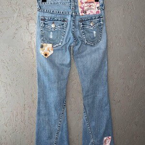 Puede incluir: Vaqueros de mezclilla azul claro con dise&ntilde;o de pernera acampanada. Los vaqueros presentan detalles desgastados, parches decorativos con motivos florales y bolsillos traseros. La etiqueta True Religion es visible en la parte trasera.