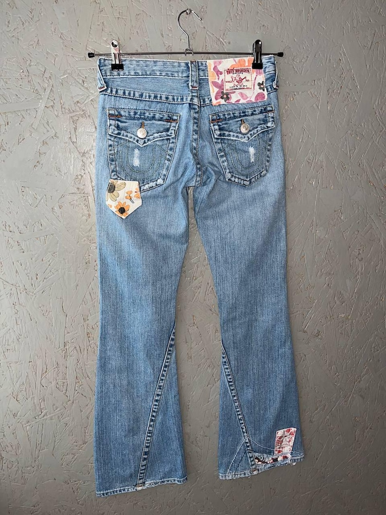 Puede incluir: Vaqueros de mezclilla azul claro con dise&ntilde;o de pernera acampanada. Los vaqueros presentan detalles desgastados, parches decorativos con motivos florales y bolsillos traseros. La etiqueta True Religion es visible en la parte trasera.