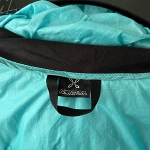 May include: Close-up of a turquoise jacket interior with a black collar and a black label. The label features the brand name "MONTURA" in white lettering, along with a stylized logo. The jacket's fabric has a slightly wrinkled texture.