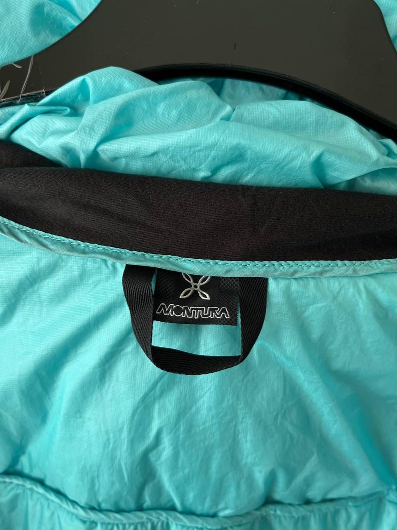 May include: Close-up of a turquoise jacket interior with a black collar and a black label. The label features the brand name "MONTURA" in white lettering, along with a stylized logo. The jacket's fabric has a slightly wrinkled texture.