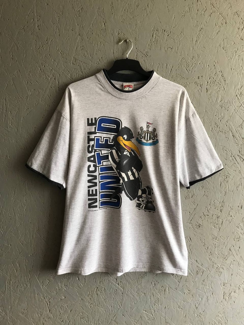 Puede incluir: Una camiseta gris con un p&aacute;jaro de dibujos animados en blanco y negro que lleva un uniforme de f&uacute;tbol. La camiseta tiene un logotipo azul y blanco que dice "Newcastle United".