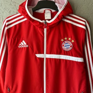 May include: Red Adidas zip-up jacket with white stripes on the sleeves and shoulders. The jacket features the FC Bayern Munich logo on the chest and the Adidas logo on the left side. It has a hood and a full-length zipper.