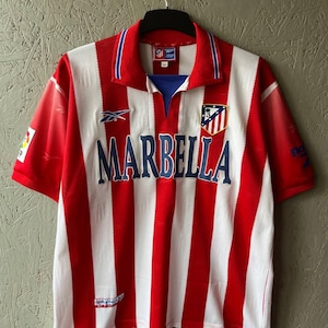 May include: Red and white striped soccer jersey with the word "MARBELLA" in blue across the front. The jersey has a blue collar and a team emblem on the chest. The Reebok logo is visible.