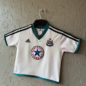 K&ouml;nnte beinhalten: Wei&szlig;es Adidas Fu&szlig;balltrikot mit schwarzen Streifen auf den Schultern und t&uuml;rkisfarbenen Verzierungen. Mit dem Newcastle Brown Ale Logo und dem Wappen von Newcastle United. Das Trikot h&auml;ngt an einem schwarzen Kleiderb&uuml;gel.