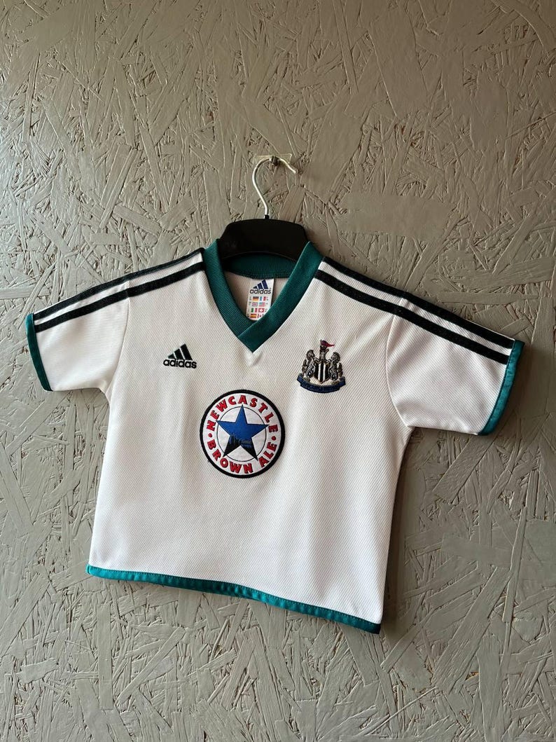 K&ouml;nnte beinhalten: Wei&szlig;es Adidas Fu&szlig;balltrikot mit schwarzen Streifen auf den Schultern und t&uuml;rkisfarbenen Verzierungen. Mit dem Newcastle Brown Ale Logo und dem Wappen von Newcastle United. Das Trikot h&auml;ngt an einem schwarzen Kleiderb&uuml;gel.