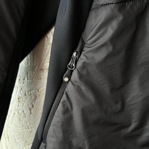 May include: Close-up of a black jacket with a zip closure. The jacket features a quilted design on the front and a smooth, solid black fabric on the sleeves. The zip pull has a small, circular logo.