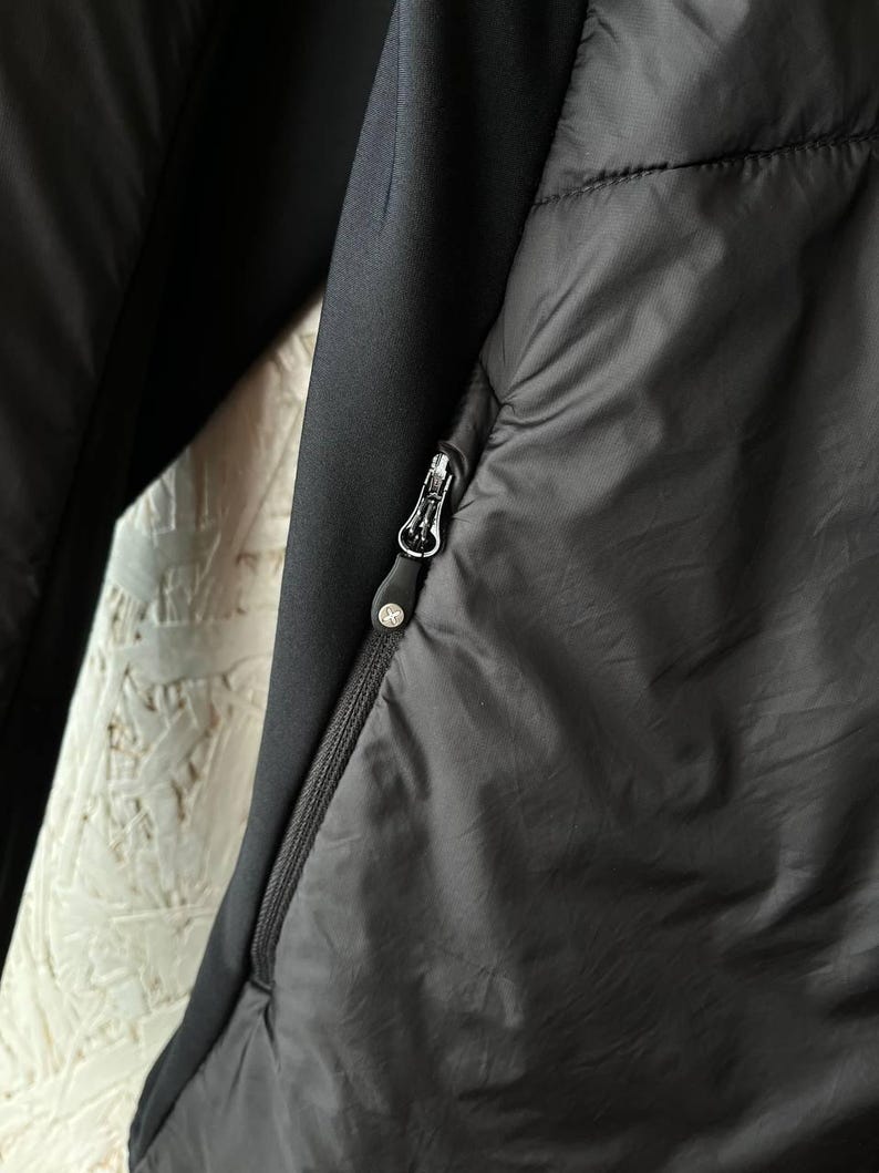 May include: Close-up of a black jacket with a zip closure. The jacket features a quilted design on the front and a smooth, solid black fabric on the sleeves. The zip pull has a small, circular logo.