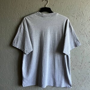 May include: A heather grey t-shirt with short sleeves. The t-shirt is hanging on a black hanger against a textured green wall.