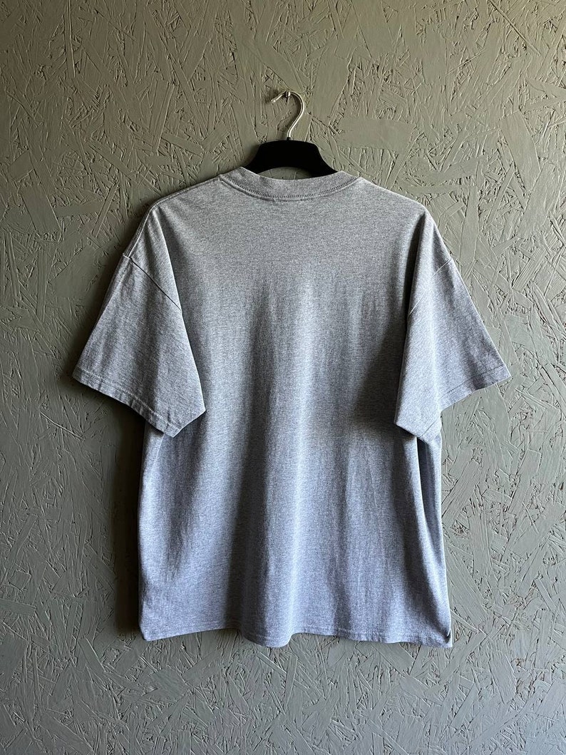 May include: A heather grey t-shirt with short sleeves. The t-shirt is hanging on a black hanger against a textured green wall.