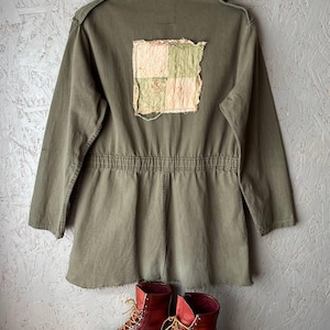 May include: An olive green utility jacket featuring a square, frayed-edge patchwork appliqué on the back in shades of cream and sage green. The jacket has long sleeves, a cinched waist, and a raw hem. Below are two brown leather lace-up boots.