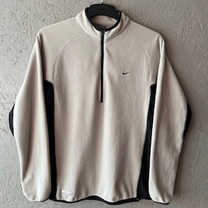 May include: Beige fleece pullover with a black zip and side panels. Features a small black Nike swoosh logo on the chest. The collar and hem are also black. The garment is hanging on a black hanger.