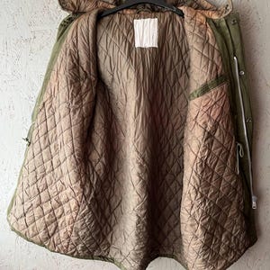 K&ouml;nnte beinhalten: Eine olivgr&uuml;ne Parka-Jacke mit braunem, gestepptem Innenfutter. Die Jacke verf&uuml;gt &uuml;ber eine Kapuze, einen durchgehenden silberfarbenen Metallrei&szlig;verschluss und ein leeres, wei&szlig;es, rechteckiges Etikett in der Mitte des inneren Halsbereichs.