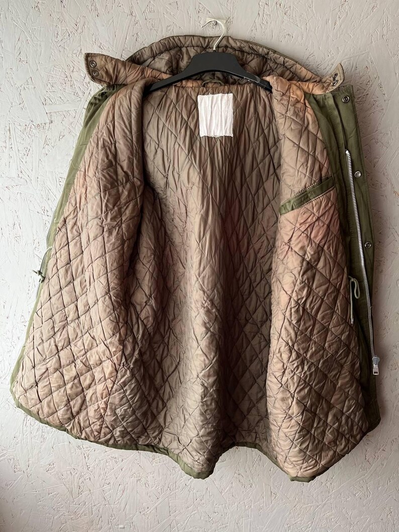 K&ouml;nnte beinhalten: Eine olivgr&uuml;ne Parka-Jacke mit braunem, gestepptem Innenfutter. Die Jacke verf&uuml;gt &uuml;ber eine Kapuze, einen durchgehenden silberfarbenen Metallrei&szlig;verschluss und ein leeres, wei&szlig;es, rechteckiges Etikett in der Mitte des inneren Halsbereichs.