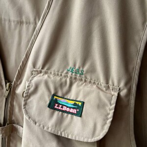 May include: A tan utility vest featuring a front zipper closure and a large pocket on the chest. The name JESS is embroidered in green thread above the pocket, which displays a rectangular patch with the LLBean logo and a landscape graphic.
