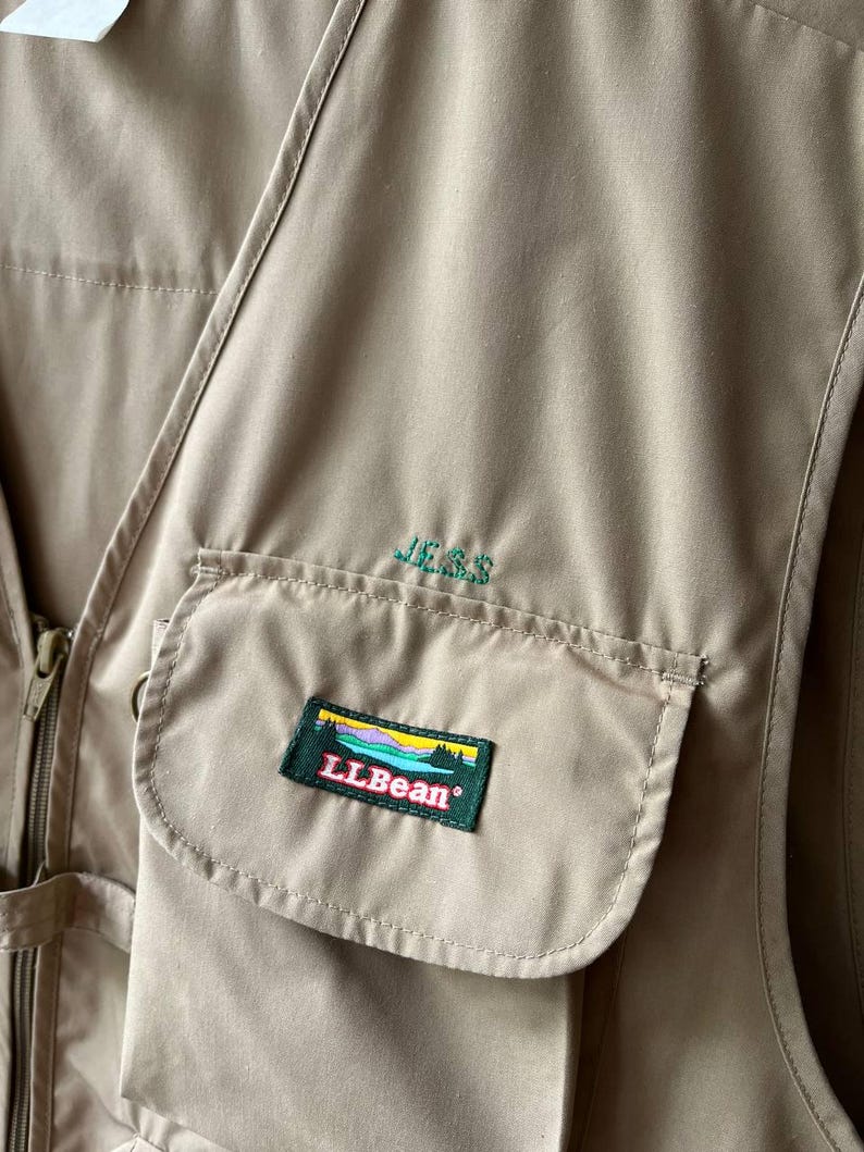 May include: A tan utility vest featuring a front zipper closure and a large pocket on the chest. The name JESS is embroidered in green thread above the pocket, which displays a rectangular patch with the LLBean logo and a landscape graphic.