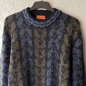 May include: A patterned sweater with a high collar in black, olive green, and blue. The label reads "MISSONI". The sweater is suspended from a black hanger. The garment is made of a textured material.
